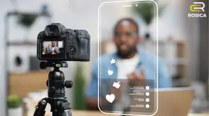 Camera on a tripod filming a man, with a translucent smartphone outline and social icons (likes, comments) overlaid; ROSICA logo in corner.