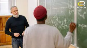 Instructor with a gray beard explains math to a student at a chalkboard filled with equations; Rosica logo upper right.