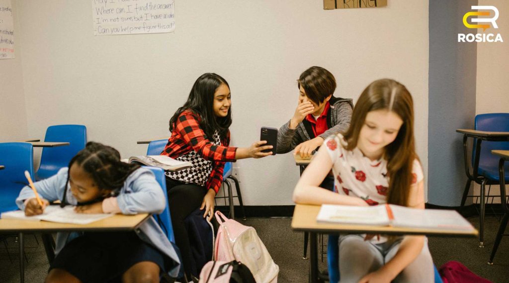 Students in a classroom laughing at image on smartphone.